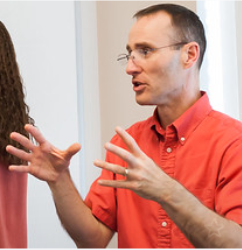 Librarian Zachary Grant wears an orange button-down shirt and speaks with his hands outstretched in a classroom.